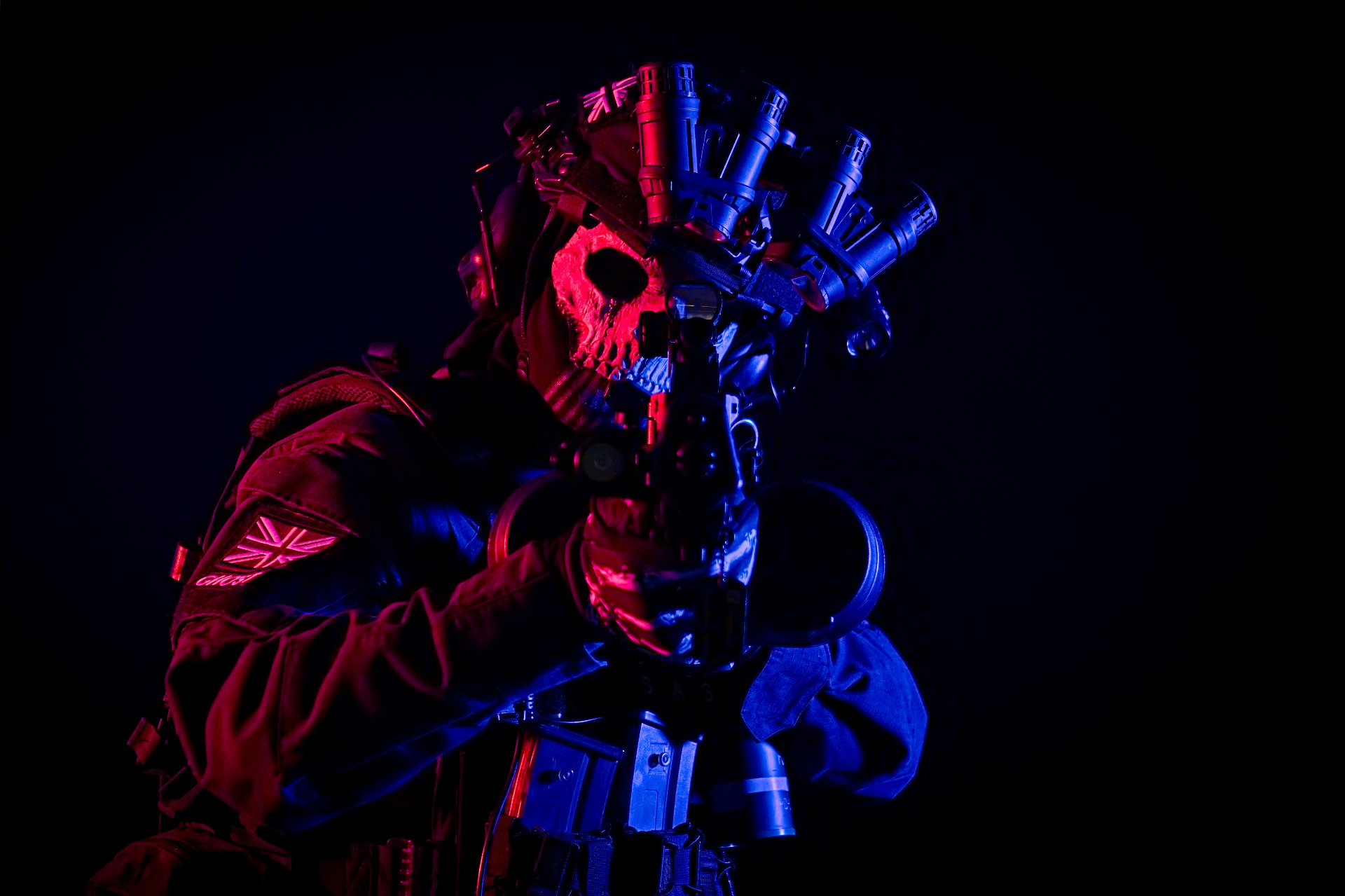 deamoon_csoplay aka calsing.deamoon v cosplay ghosta z taktické herní série. Má helmu s nočním vizorem, masku ve tvaru lebky a replikou zbraně míří na diváka. Vystupuje z tmavého pozadí osvícen červeným světlem z levé strany a modrým světlem z pravé strany.