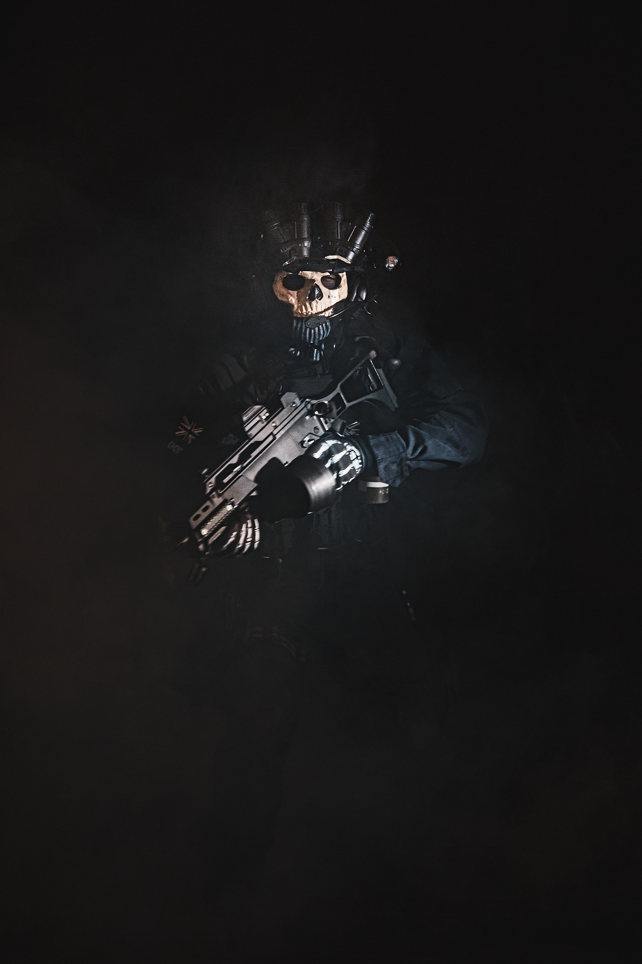 Deamoon_cosplay vystupující ze tmy a kouře v taktickém oblečení se zbraní v ruce. Jedná se o csoplay postavy Ghost z taktické herní série, s maskou výrazné struktury lebky, nočním vizorem a helmou.