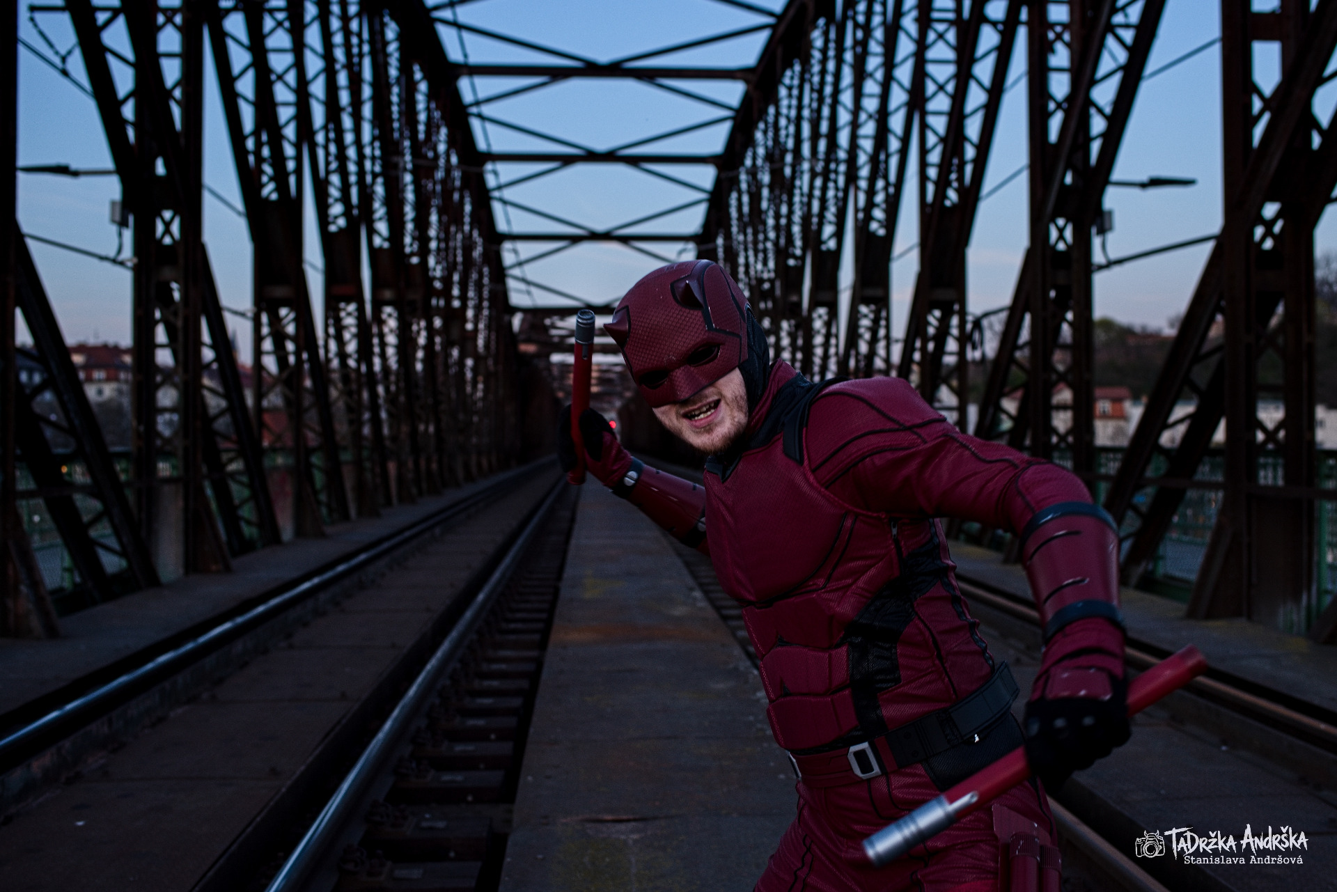 Cosplay daredevila v červeném kostýmu. Model je v bojové póze, napřažená rozložené slepecká hůl v útoku proti divákovi. V pozadí se rýsuje industriální pozadí Prahy