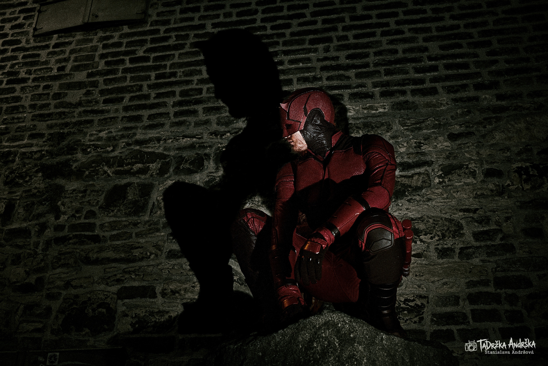 sattler.jan jako cosplay daredevila v noční Praze. Dřepí na velkém kameni, připraven vyrazit do akce, osvětluje ho ostré světlo, které na kamenné zdi vytváří ostré stíny jeho siluety.
Autorem fotografie je Tadržka Andrška